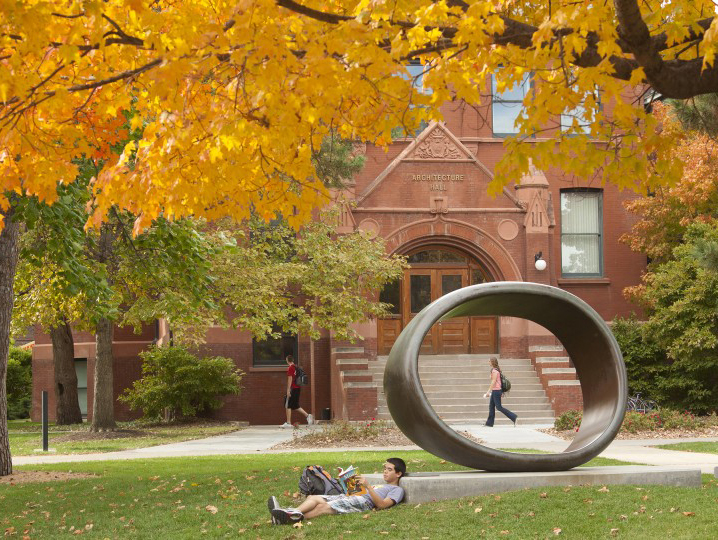 A male student reclines and reads a book in the Sheldon Museum of Art's Sculpture Garden under the golden colors of the autumn tree foliage.