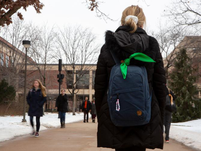 A female student walks across campus with a green bandana tied on her backpack.