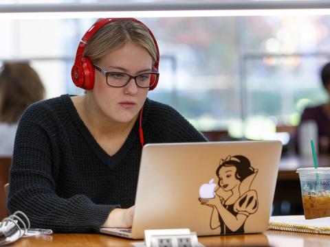 Female students wears headphones and looks at her laptop screen.