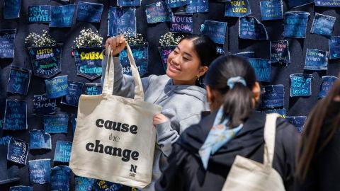 Student takes a photo with a Cause The Change tote bag in front of a denim wall in support of sexual assault survivors on Denim Day 2023.