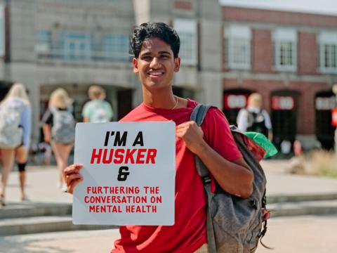 Naren smiles for a photo holding a sign that says "I'm a Husker & furthering the conversation on mental health."